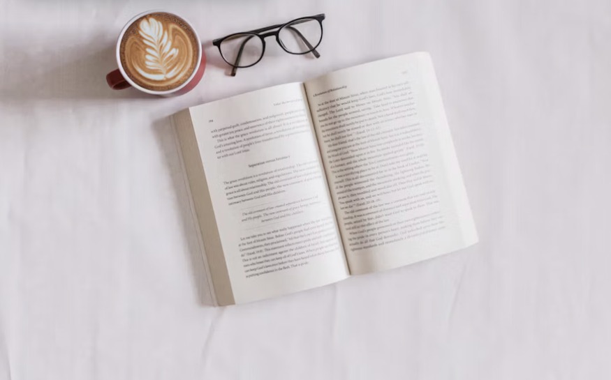Un livre ouvert, une tasse de café et une paire de lunettes posés sur un drap clair